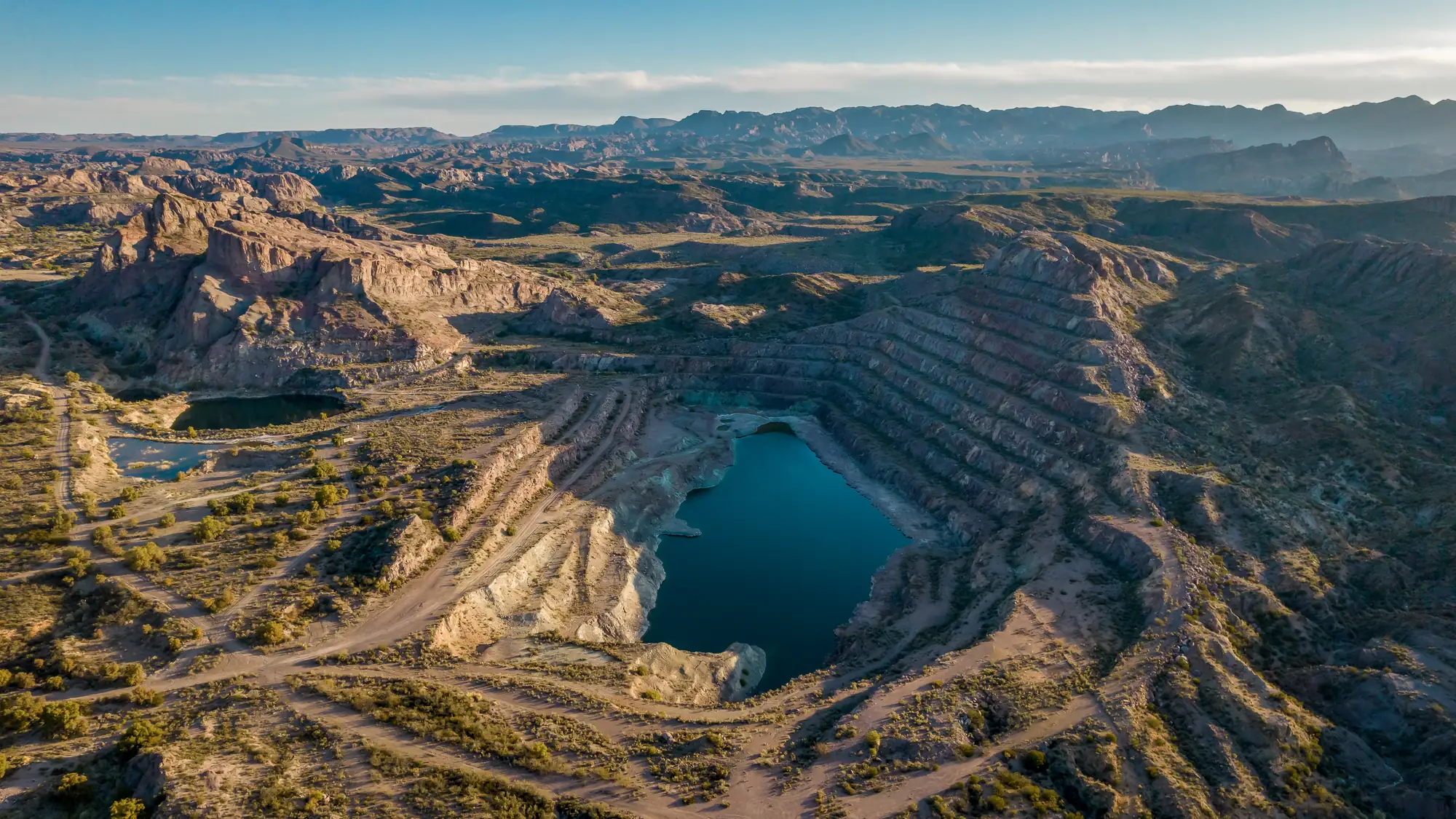 Aerial view of a terraced open pit holding a deep blue lake, desert ranges beyond